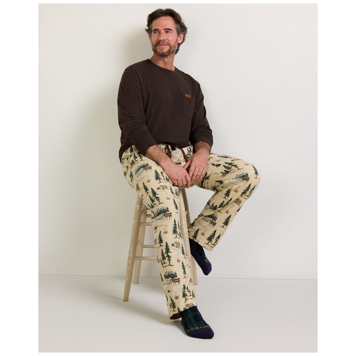 Man seated on stool wearing Pendleton tan camping-print pajama pants with brown top