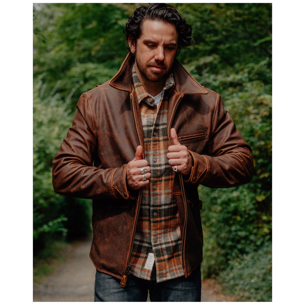 Man wearing brown distressed bison leather delivery jacket over plaid flannel shirt outdoors